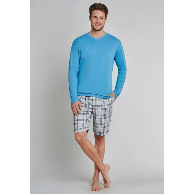 Model in blauw Schiesser heren nachtshirt 163846 met lange mouwen en V-hals, gecombineerd met geruite shorts. - Jambelles